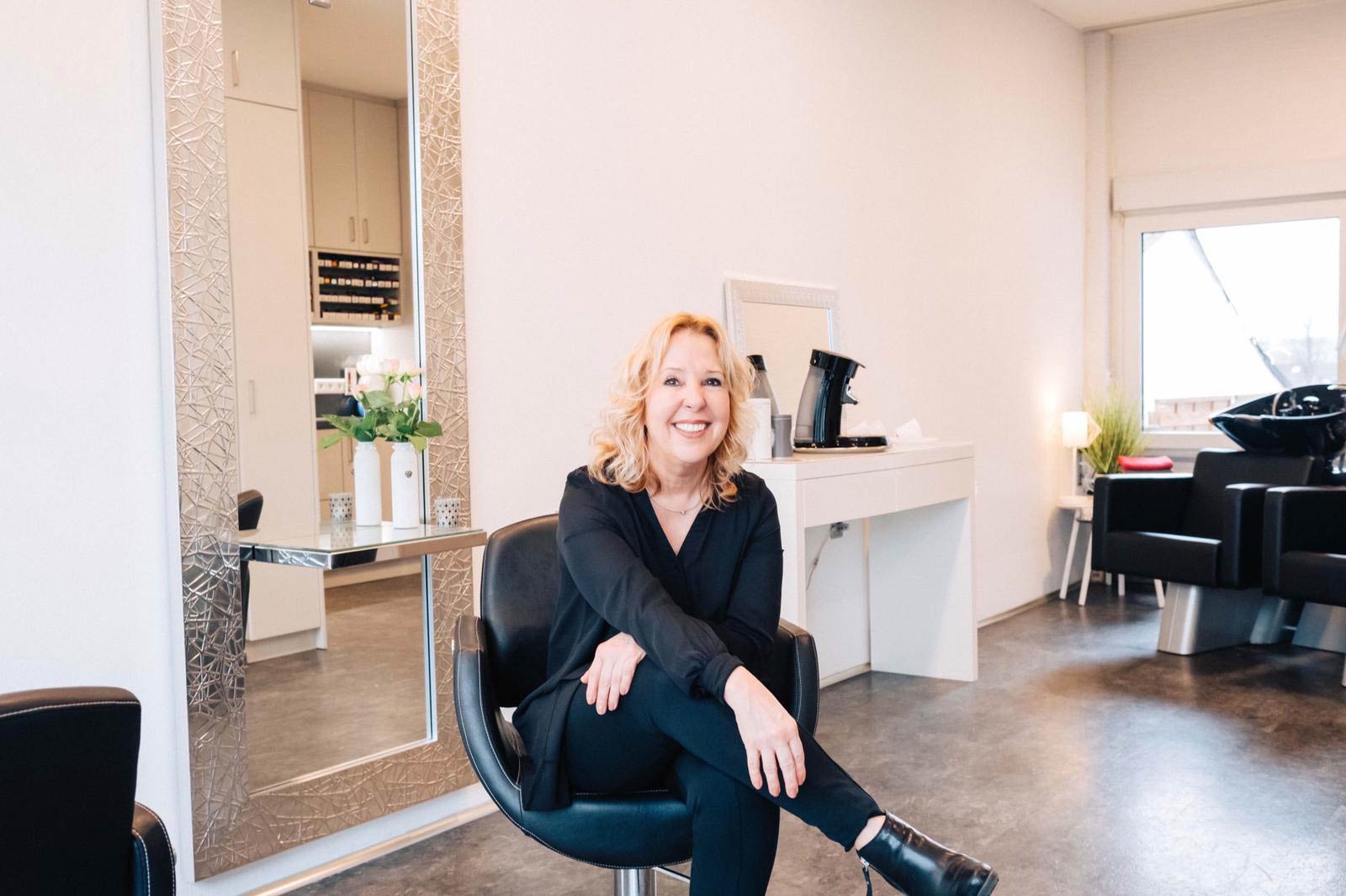 Christiane Franz sitzt lächelnd auf einem Friseurstuhl in ihrem Salon, hinter ihr ein silberner Spiegel