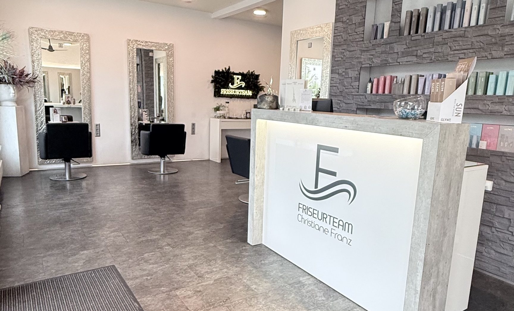 Blick in den Salon: Empfangstresen mit Logo vor der grauen Steinwand, dahinter Bedienplätze mit silbern gerahmten Spiegeln